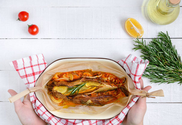 Hands hold freshly cooked fish, vegetables, aromatic herbs, olive oil on a white wooden table. Top view with copy space, flat lay. Food background. - Valokuva, kuva