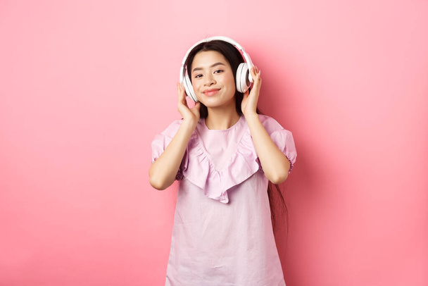 Güzel Asyalı kadın, kablosuz kulaklıkla müzik dinliyor, kameraya gülümsüyor, pembe arka planda elbise giymiş. - Fotoğraf, Görsel