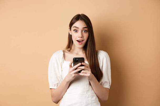 Отличные новости по телефону. Удивительная молодая женщина сказать ничего себе после прочтения онлайн объявление на смартфоне, стоя на бежевом фоне - Фото, изображение
