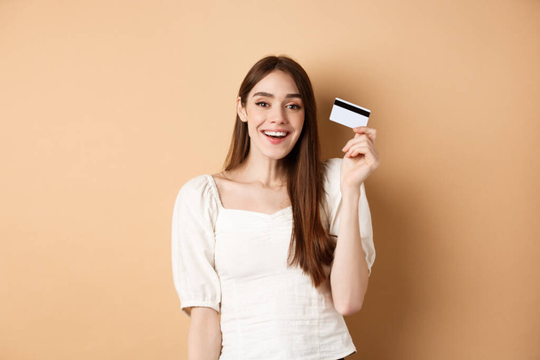 Jovem alegre tem seu cartão de crédito de plástico e sorrindo satisfeito, de pé satisfeito com fundo bege - Foto, Imagem