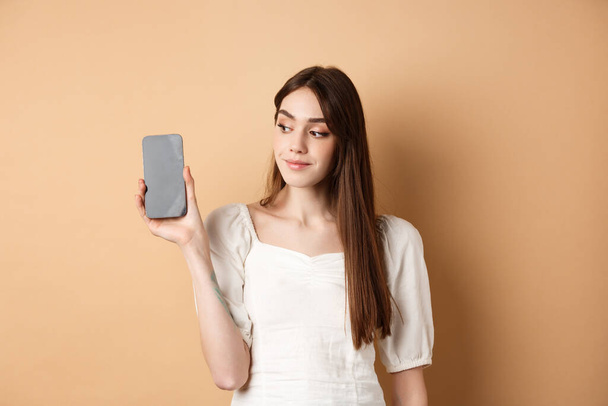 Приваблива молода жінка, показуючи порожній екран смартфона, посміхаючись і озираючись вбік, стоїть на бежевому фоні
 - Фото, зображення