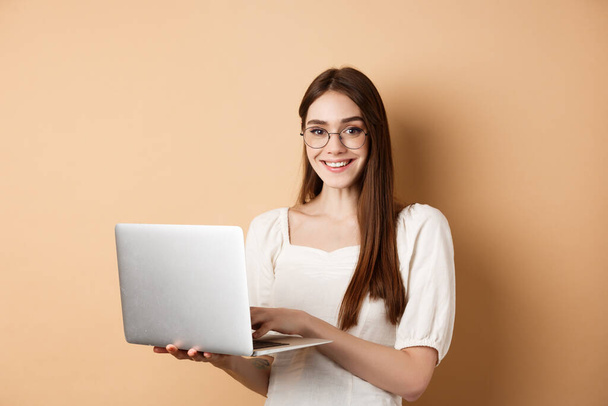 Studentessa sorridente che lavora sul computer portatile, indossa occhiali e sembra felice, utilizzando il computer mentre in piedi sullo sfondo beige - Foto, immagini