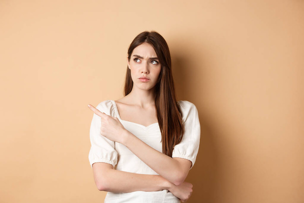 Suspicious woman in dress looking, pointing left, frowning and staring with disbelief and doubts, standing on beige background - Photo, Image