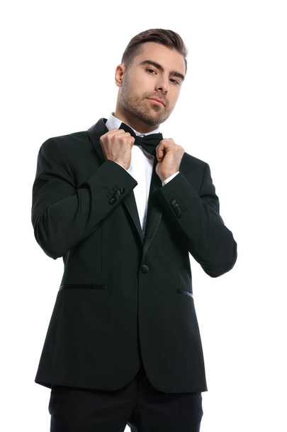 elegante jovem empresário em preto smoking fixação bowtie e posando isolado no fundo branco no estúdio, retrato - Foto, Imagem