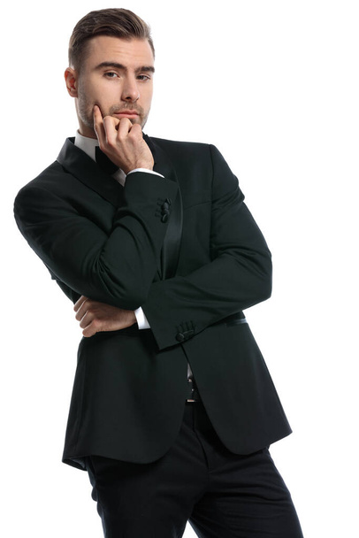 portret troskliwego biznesmena w czarnym smokingu, trzymającego się za brodę i myślącego, stojącego i pozującego na białym tle w studio - Zdjęcie, obraz