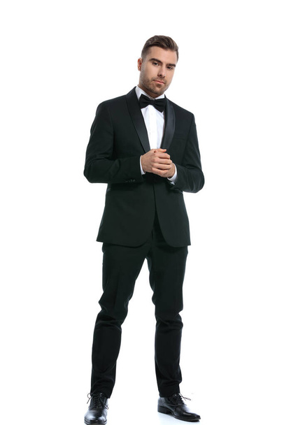 full body picture of elegant young businessman in black tuxedo holding hands and posing isolated on white background in studio - Φωτογραφία, εικόνα