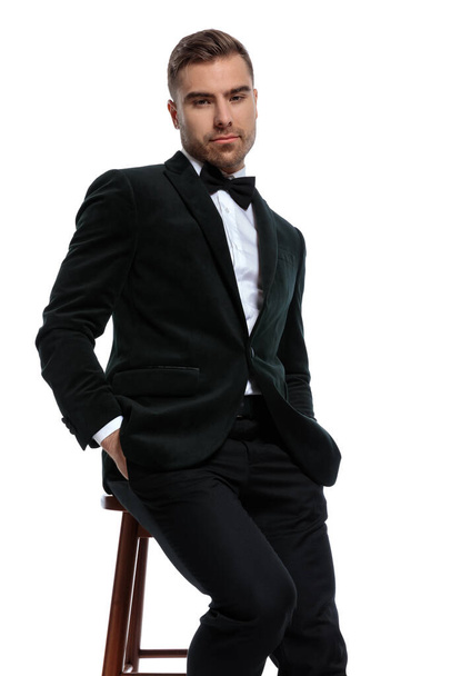 elegant young businessman in black tuxedo holding hands in pockets and sitting isolated on white background in studio - Foto, afbeelding