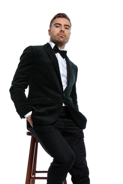 seated unshaved businessman in black tuxedo holding hands in pockets and confidently posing on white background in studio - Fotó, kép