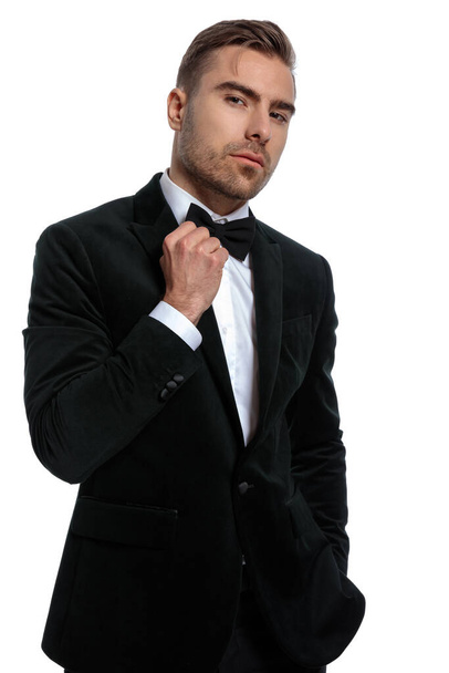 confiante cara elegante em smoking preto segurando mão no bolso e ajustando bowtie enquanto posando isolado no fundo branco no estúdio - Foto, Imagem