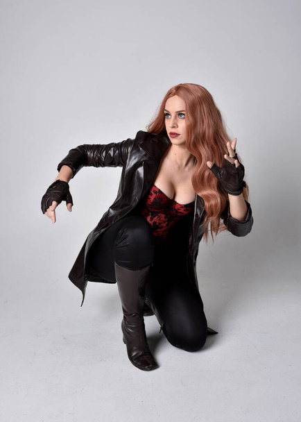 full length portrait of girl with long red hair wearing dark leather coat, corset and boots. Sitting pose facing front on with  magical hand gestures against a studio background. - Foto, Bild