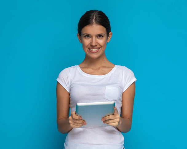 jong casual vrouw houden van een tablet in haar handen en glimlachen naar de camera tegen een blauwe achtergrond - Foto, afbeelding