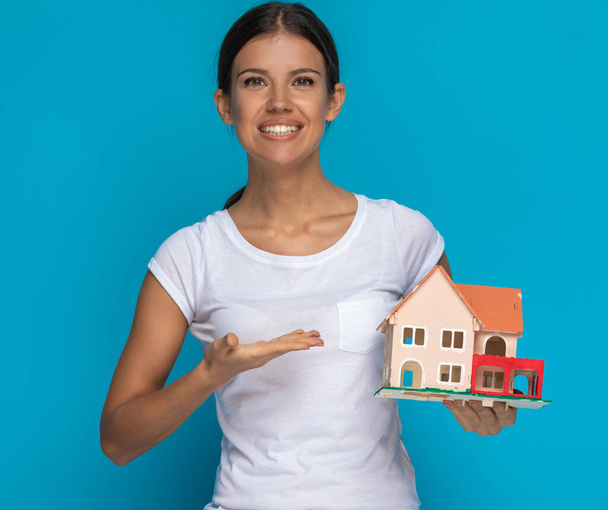 περιστασιακή γυναίκα παρουσιάζει ένα μοντέλο σπιτιού με ένα χαμόγελο στο πρόσωπό της σε μπλε φόντο - Φωτογραφία, εικόνα