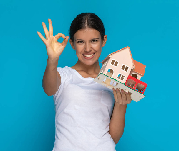 casual woman holding a house in her hand and giving a thumbs up, feeling excited against blue background - Photo, Image