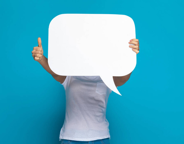casual woman hiding behind a speech bubble and giving a thumbs up against blue background - Foto, Imagen