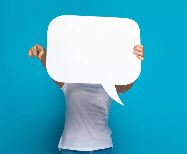 casual woman hiding behing a speech bubble and pointing at the camera against blue background - Photo, Image