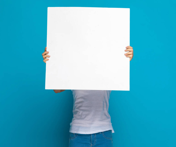 περιστασιακή γυναίκα κρύβει το πρόσωπό της πίσω από μια λευκή πινακίδα σε λευκό φόντο στούντιο - Φωτογραφία, εικόνα