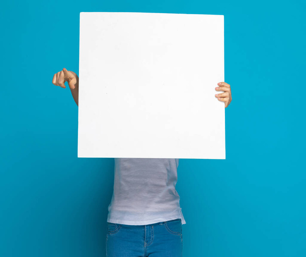 casual woman hiding behind a blank billboard and pointing at the camera against blue background - Foto, afbeelding