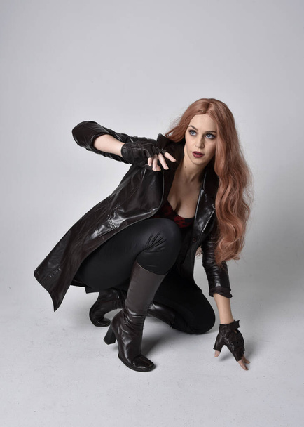 full length portrait of girl with long red hair wearing dark leather coat, corset and boots. Sitting pose facing front on with  magical hand gestures against a studio background. - Fotó, kép