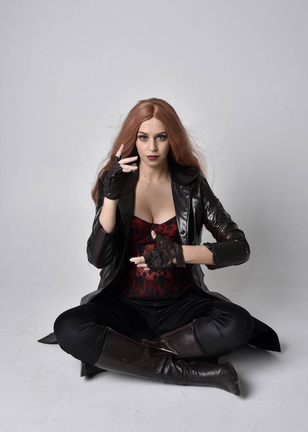 full length portrait of girl with long red hair wearing dark leather coat, corset and boots. Sitting pose facing front on with  magical hand gestures against a studio background. - Photo, Image