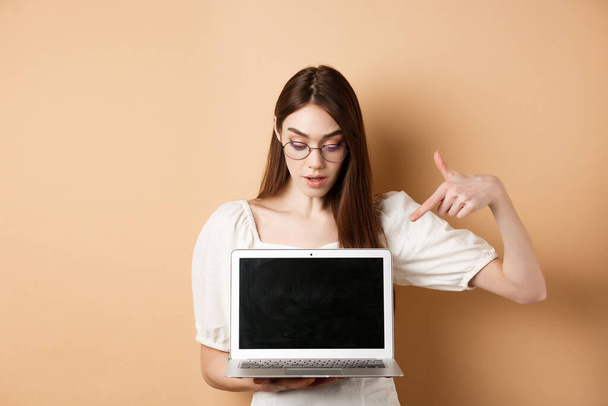 Περίεργη κοπέλα με γυαλιά που δείχνει την οθόνη του φορητού υπολογιστή, ελέγξτε online deal, επιδείξτε το έργο στον υπολογιστή, στέκεται σε μπεζ φόντο - Φωτογραφία, εικόνα