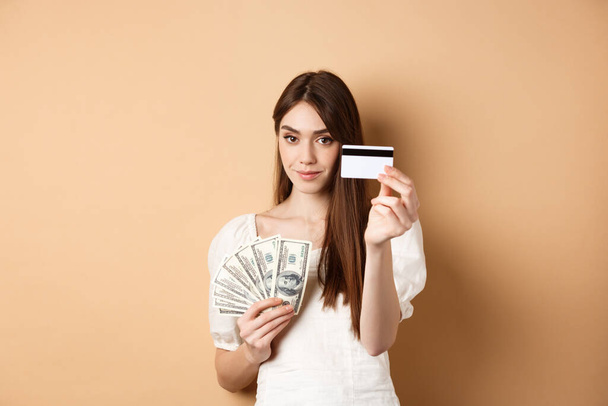 Plastik kredi kartı gösteren genç bir kadın, dolar yerine temassız ödemeyi tercih ediyor, bej arka planda duruyor. - Fotoğraf, Görsel