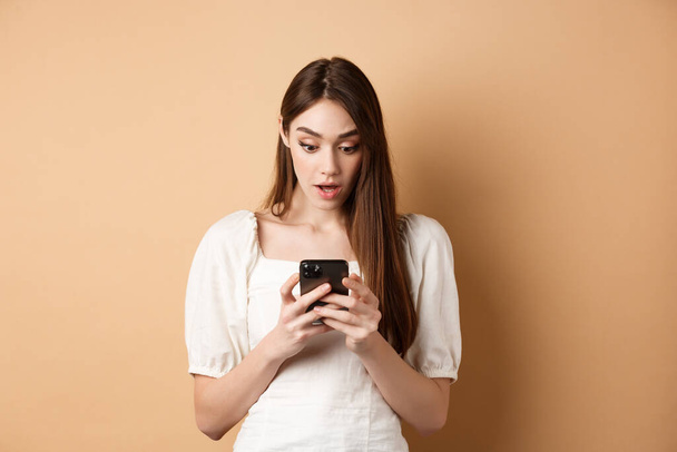 Frau blickt überrascht auf Smartphone-Bildschirm, liest schockierende Nachricht auf Handy, steht erstaunt auf beigem Hintergrund - Foto, Bild