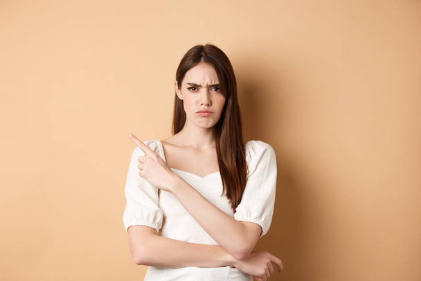 Angry frowning girl pointing left at empty space, looking disappointed and upset, scolding you or accuse, standing on beige background - Fotoğraf, Görsel