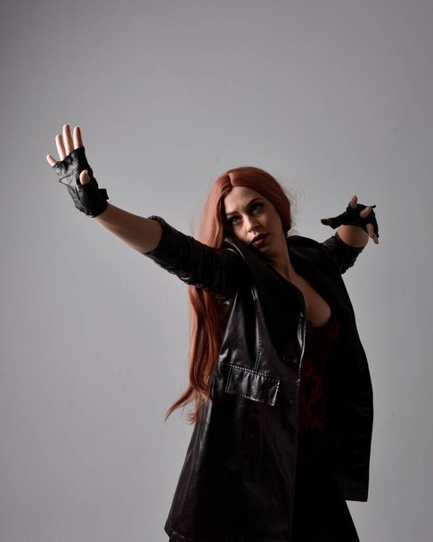 Κοντινό πορτρέτο του κοριτσιού με μακριά κόκκινα μαλλιά φορώντας σκούρο δερμάτινο παλτό, κορσέ και γάντια. Τοποθέτηση με χειρονομίες χειρονομίες σαν να ρίχνει ξόρκι. Moody πίσω φωτισμένο φωτισμό σε φόντο στούντιο. - Φωτογραφία, εικόνα