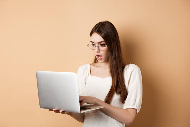 Bezorgd meisje kijken naar laptop scherm, typen op de computer met gericht gezicht. Vrouw freelancer werken in internet, staan op beige achtergrond - Foto, afbeelding