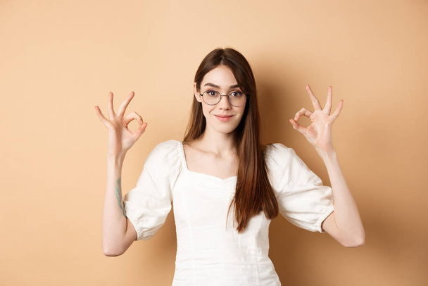 Ικανοποιημένο θηλυκό μοντέλο σε γυαλιά δείχνουν εντάξει σημάδι, αναζητούν ευχαριστημένοι, συμφωνούν και εγκρίνουν, στέκεται σε μπεζ φόντο - Φωτογραφία, εικόνα