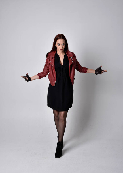Full length portrait of girl wearing red leather jacket, tights with boots and gloves. Posing with gestural hand movements as if casting spell against a studio background. - Φωτογραφία, εικόνα