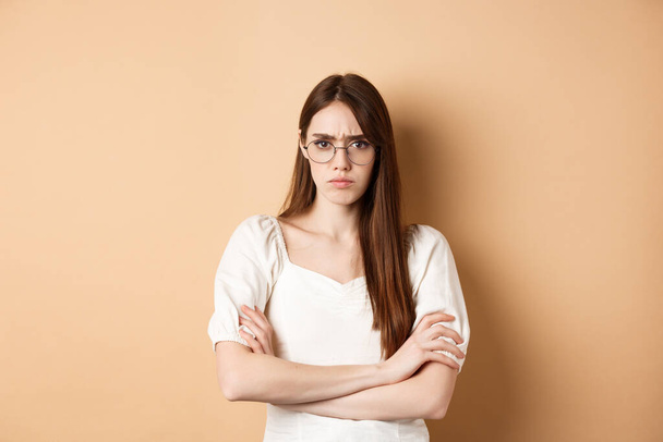 Angry and offended girl in glasses frowning, cross arms on chest and sulking, standing defensive on beige background - Foto, Bild