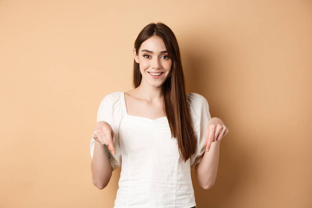 Cheerful young girl with lovely smile, pointing fingers down, making announcement or inviting click link, showing logo, beige background - Photo, Image