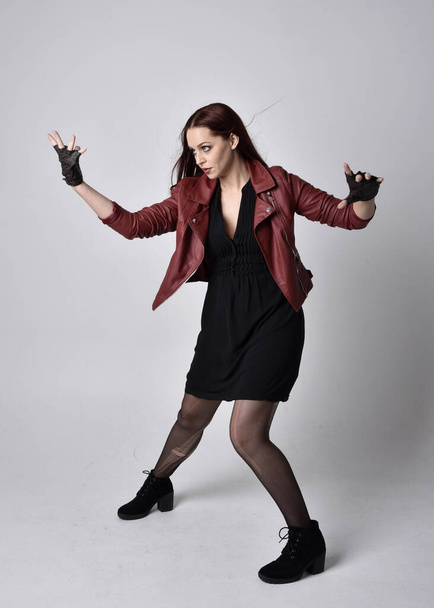 Volledig portret van meisje in rood leren jasje, panty met laarzen en handschoenen. Poseren met gebaar handbewegingen alsof casting spell tegen een studio achtergrond. - Foto, afbeelding