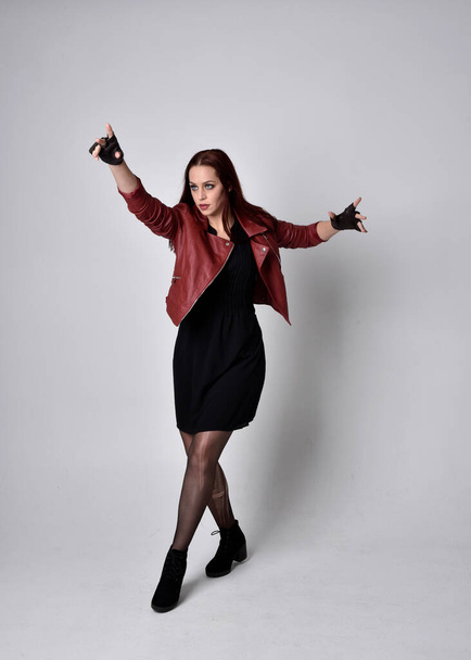 Full length portrait of girl wearing red leather jacket, tights with boots and gloves. Posing with gestural hand movements as if casting spell against a studio background. - Photo, image