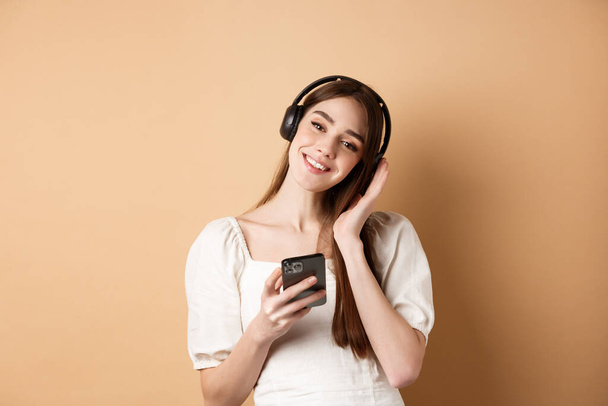 Roztomilé usmívající se dívka poslech hudby v bezdrátových sluchátkách, pomocí smartphonu, stojící na béžovém pozadí - Fotografie, Obrázek