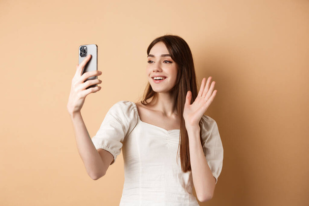 Meisje video chat op telefoon, afzien van de hand op mobiele camera en zeggen hallo, praten met een vriend, glimlachen en staan op beige achtergrond - Foto, afbeelding