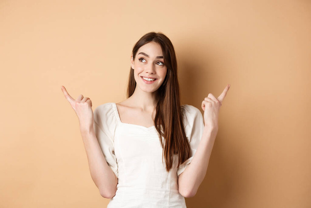 Dreamy caucasian girl making choice, pointing fingers sideways and looking at upper left corner pensive, picking product, standing on beige background - Fotó, kép