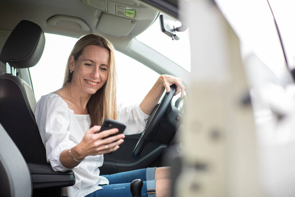Pretty middle aged woman having at the wheel of her car, using a smartphone - Foto, Imagem