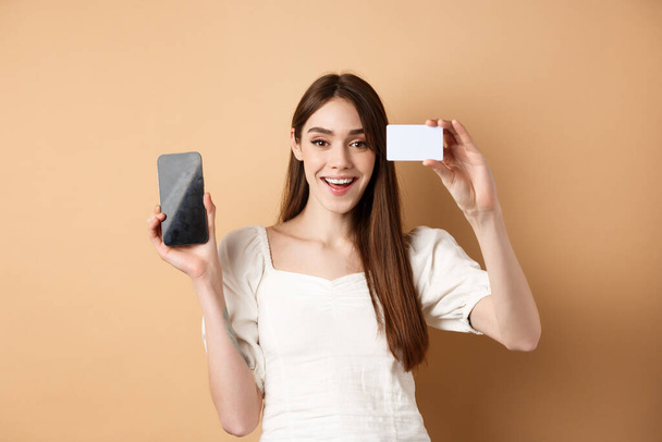 Giovane donna allegra che mostra carta di credito di plastica e schermo vuoto del telefono cellulare, sorridente soddisfatto della fotocamera, in piedi su sfondo beige - Foto, immagini