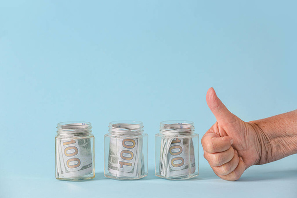 Hand of senior woman showing thumb-up gesture near jars with money on color background. Concept of pension - Photo, Image