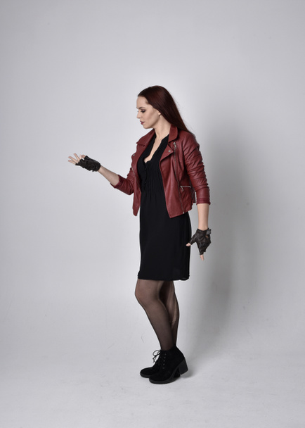 Портрет девушки в красной кожаной куртке, колготки с сапогами и перчатками. Позирование с жестикулирующими движениями рук, как будто накладывая заклинание на студийный фон. - Фото, изображение
