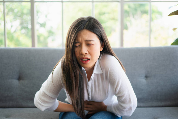 Mujer asiática infeliz sentada en el sofá y sosteniendo el sufrimiento del estómago. Dolor abdominal que proviene de la menstruación, diarrea o indigestión. Concepto de enfermedad y asistencia sanitaria - Foto, imagen