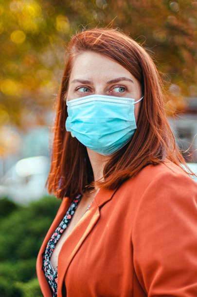 20 yaşında, koruyucu bir sağlık maskesi takmış beyaz bir kadın sonbahar parkında gözlerini kaçırıyor. Coronavirüs salgını sırasında kamusal alanda güvenlik. - Fotoğraf, Görsel
