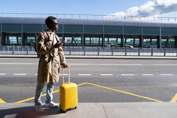 Black traveler man with suitcase stands in airport terminal holding phone calling looking for a taxi - Foto, immagini