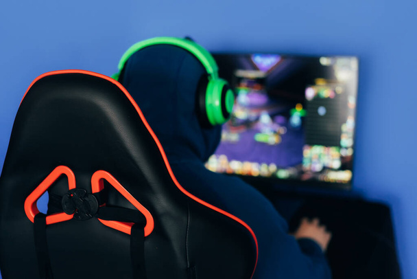 The gamer sits on a gaming chair and plays computer games. The player has green headphones on his head. - Photo, Image