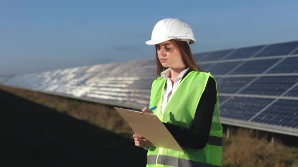 Egy női mérnök vizsgálja a napelemeket. Adatokat visz be a dokumentumba. Zöld mellény és kemény kalap van rajta. Napelemek a háttérben. 4k - Felvétel, videó