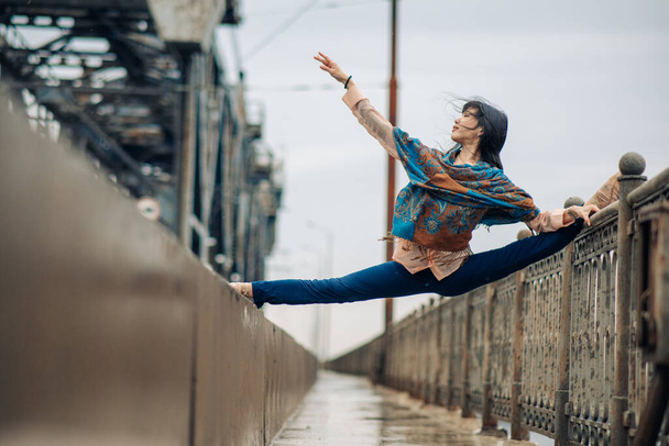 Japanese ballerina sitting in twine pose on the bridge against the background of sidewalk and railings. - Φωτογραφία, εικόνα