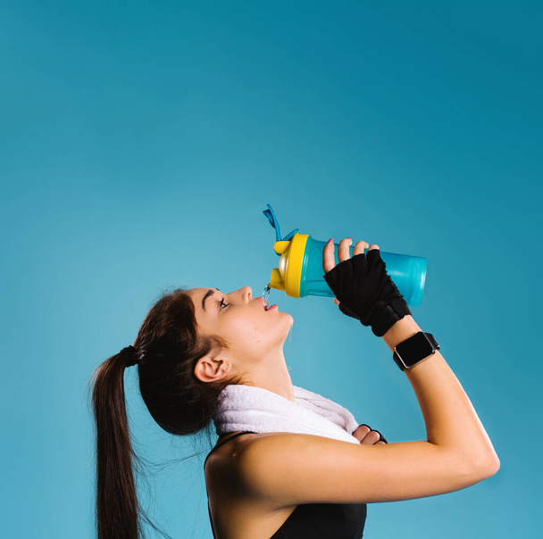 Sportlerin mit Handtuch um den Hals nach dem Training mit Durstgetränken auf blauem Grund. Leere obere Fläche für Werbung. - Foto, Bild
