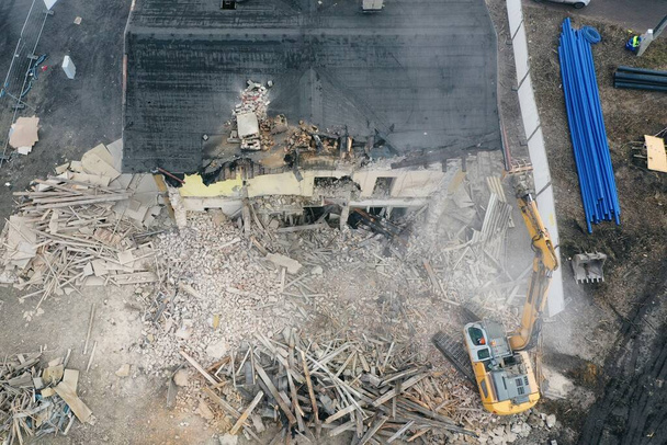 Ο κίτρινος εκσκαφέας καταστρέφει κτίριο. Η βαριά μηχανή κατεδαφίζει ένα κτίριο από τούβλα. Κατεδάφιση του κτιρίου. Κατασκευαστικές εργασίες κατεδάφισης εναέρια προβολή φωτογραφιών drone - Φωτογραφία, εικόνα